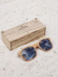 Load image into Gallery viewer, Zebrawood wooden sunglasses with black polarized lenses and a box on sandy beach.
