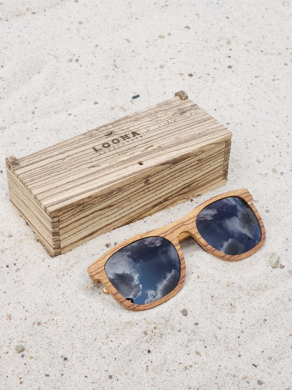 Zebrawood wooden sunglasses with black polarized lenses and a box on sandy beach.