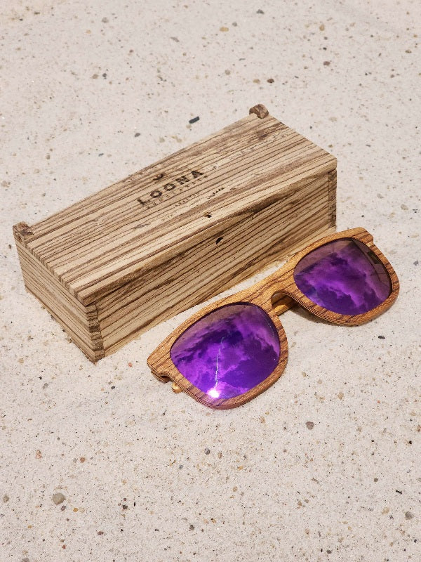 Zebrawood wooden sunglasses with purple polarized lenses and a box on sandy beach.