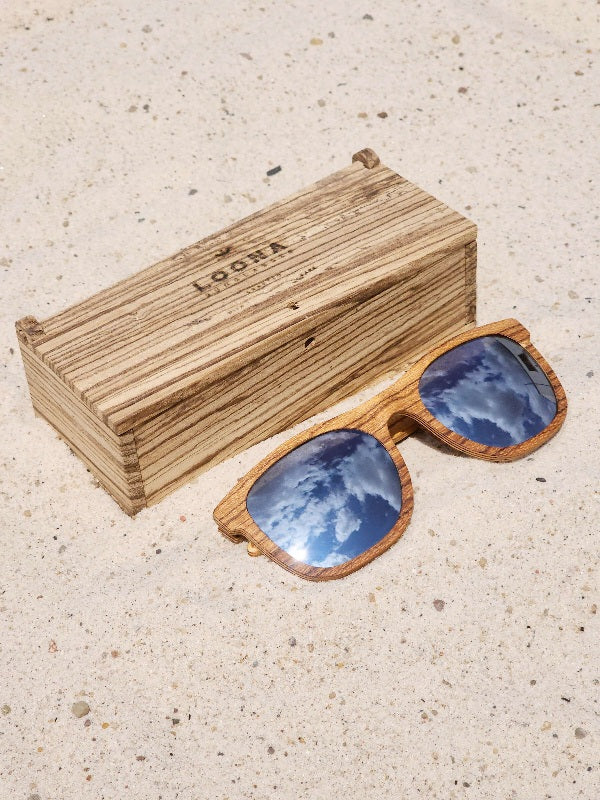 Zebrawood wooden sunglasses with silver polarized lenses and a box on sandy beach.