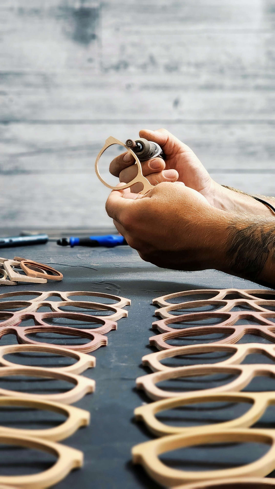 A person’s hand assembling wooden sunglasses on a table scattered with similar objects and tools.