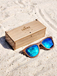 Load image into Gallery viewer, Walnut wooden sunglasses with blue polarized lenses and a box on sandy beach.
