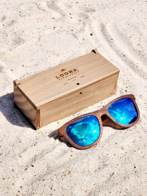 Walnut wooden sunglasses with blue polarized lenses and a box on sandy beach.