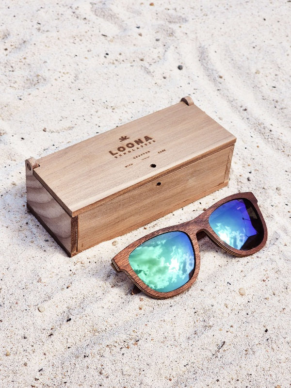 Walnut wooden sunglasses with green polarized lenses and a box on sandy beach.