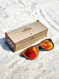Load image into Gallery viewer, Walnut wooden sunglasses with red polarized lenses and a box on sandy beach.
