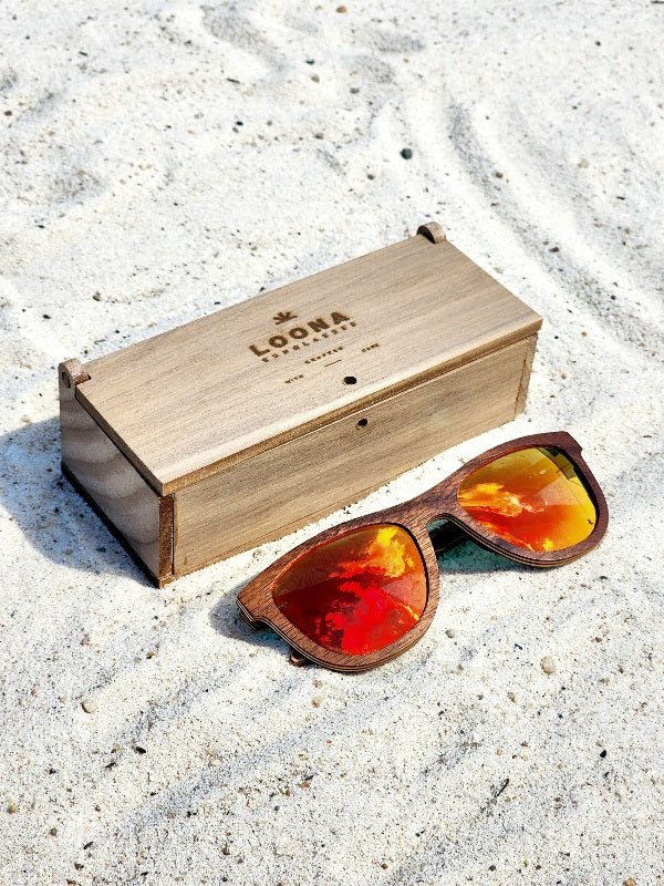 Walnut wooden sunglasses with red polarized lenses and a box on sandy beach.