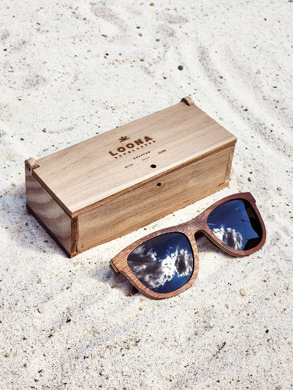 Walnut wooden sunglasses with black polarized lenses and a box on sandy beach.