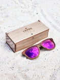 Load image into Gallery viewer, Walnut wooden sunglasses with purple polarized lenses and a box on sandy beach.
