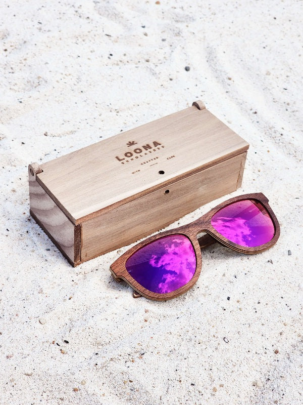 Walnut wooden sunglasses with purple polarized lenses and a box on sandy beach.