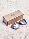 Load image into Gallery viewer, Walnut wooden sunglasses with silver polarized lenses and a box on sandy beach.
