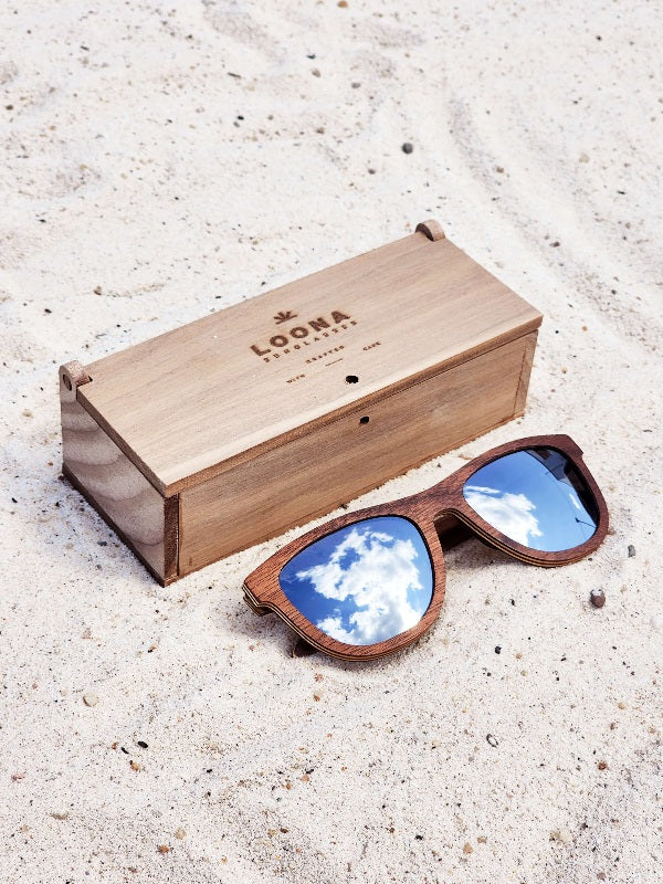 Walnut wooden sunglasses with silver polarized lenses and a box on sandy beach.