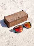 Load image into Gallery viewer, Wenge wooden sunglasses with red polarized lenses and a box on sandy beach.
