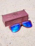 Load image into Gallery viewer, Purpleheart wooden sunglasses with blue polarized lenses and a box on sandy beach.
