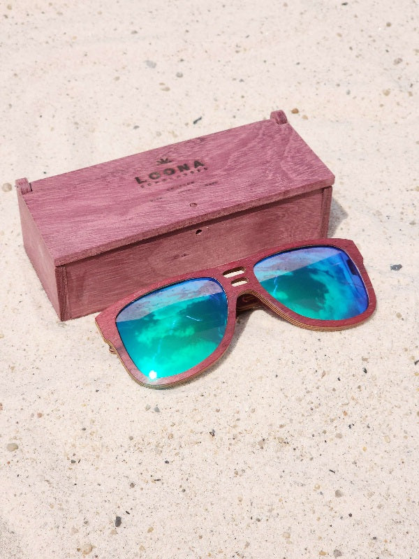 Purpleheart wooden sunglasses with green polarized lenses and a box on sandy beach.