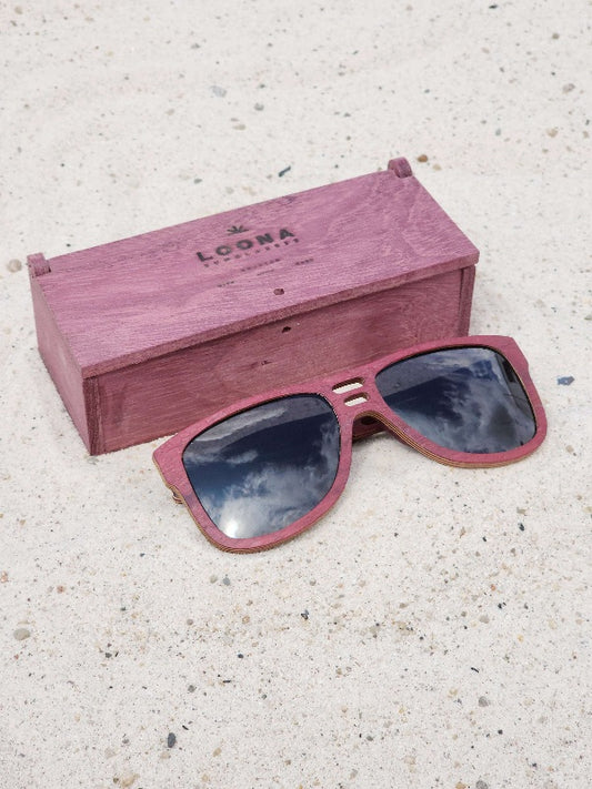 Purpleheart wooden sunglasses with black polarized lenses and a box on sandy beach.