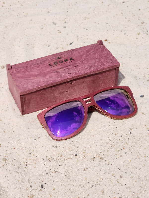 Purpleheart wooden sunglasses with purple polarized lenses and a box on sandy beach.