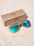 Load image into Gallery viewer, Walnut wooden sunglasses with green polarized lenses and a box on sandy beach.

