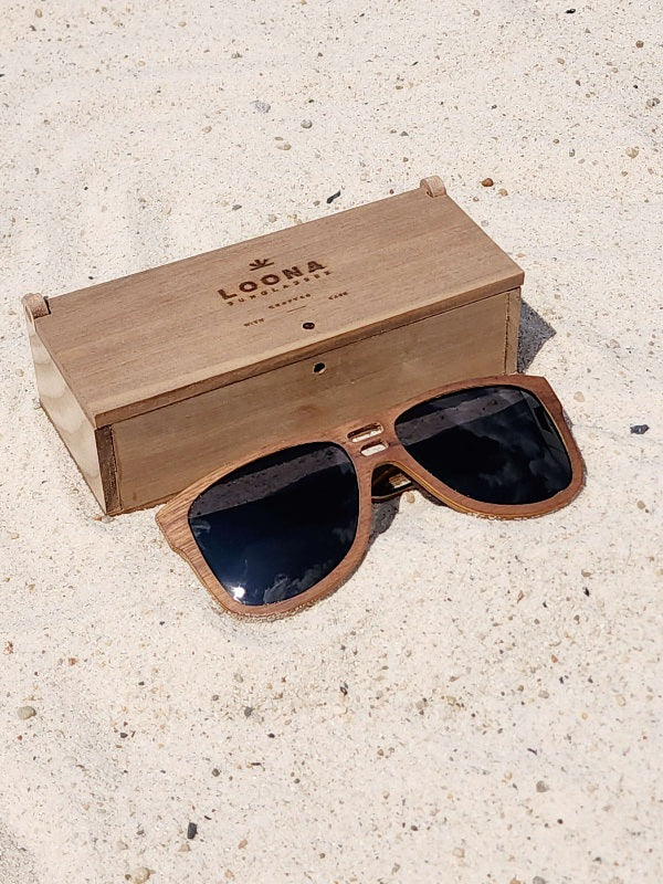 Walnut wooden sunglasses with black polarized lenses and a box on sandy beach.