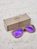 Load image into Gallery viewer, Walnut wooden sunglasses with purple polarized lenses and a box on sandy beach.

