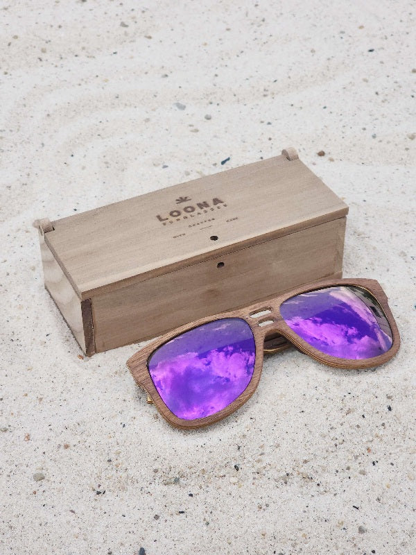 Walnut wooden sunglasses with purple polarized lenses and a box on sandy beach.