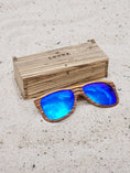 Load image into Gallery viewer, Zebrawood wooden sunglasses with blue polarized lenses and a box on sandy beach.
