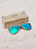 Load image into Gallery viewer, Zebrawood wooden sunglasses with green polarized lenses and a box on sandy beach.
