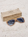 Load image into Gallery viewer, Zebrawood wooden sunglasses with black polarized lenses and a box on sandy beach.
