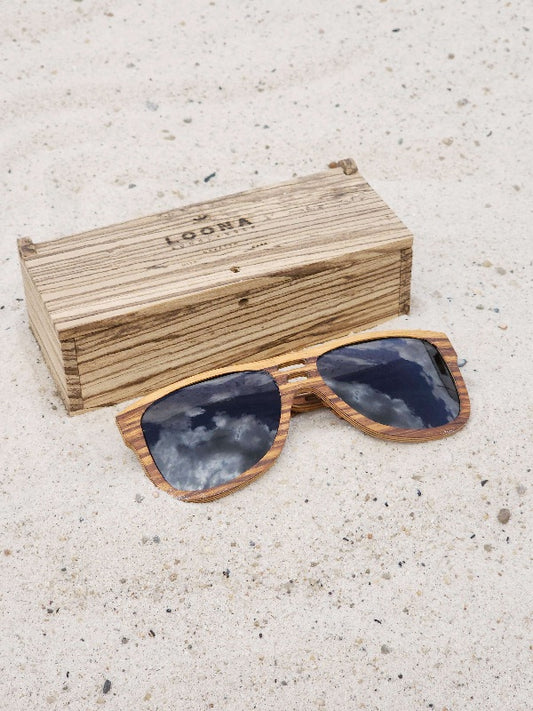 Zebrawood wooden sunglasses with black polarized lenses and a box on sandy beach.