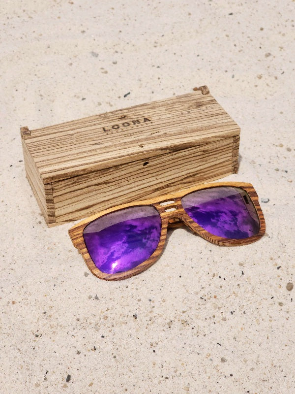 Zebrawood wooden sunglasses with purple polarized lenses and a box on sandy beach.