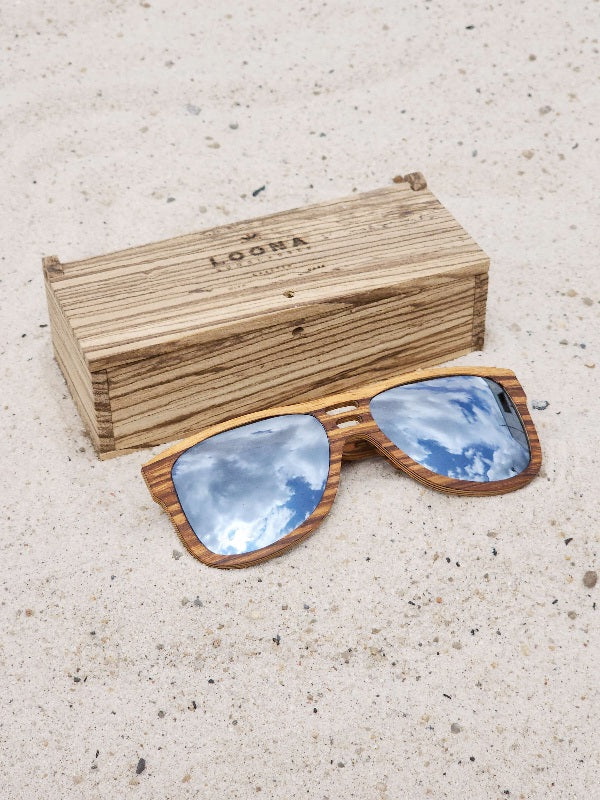 Zebrawood wooden sunglasses with silver polarized lenses and a box on sandy beach.