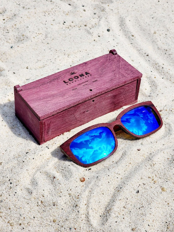 Purpleheart wooden sunglasses with blue polarized lenses and a box on sandy beach.