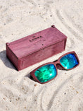 Load image into Gallery viewer, Purpleheart wooden sunglasses with green polarized lenses and a box on sandy beach.
