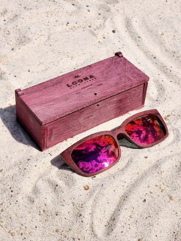 Purpleheart wooden sunglasses with red polarized lenses and a box on sandy beach.