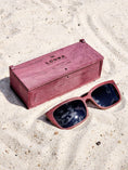 Load image into Gallery viewer, Purpleheart wooden sunglasses with black polarized lenses and a box on sandy beach.
