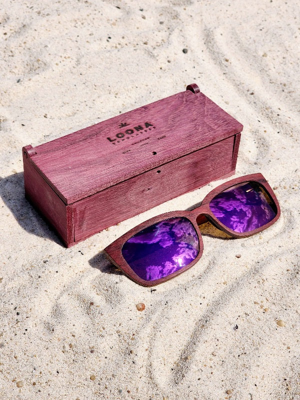 Purpleheart wooden sunglasses with purple polarized lenses and a box on sandy beach.