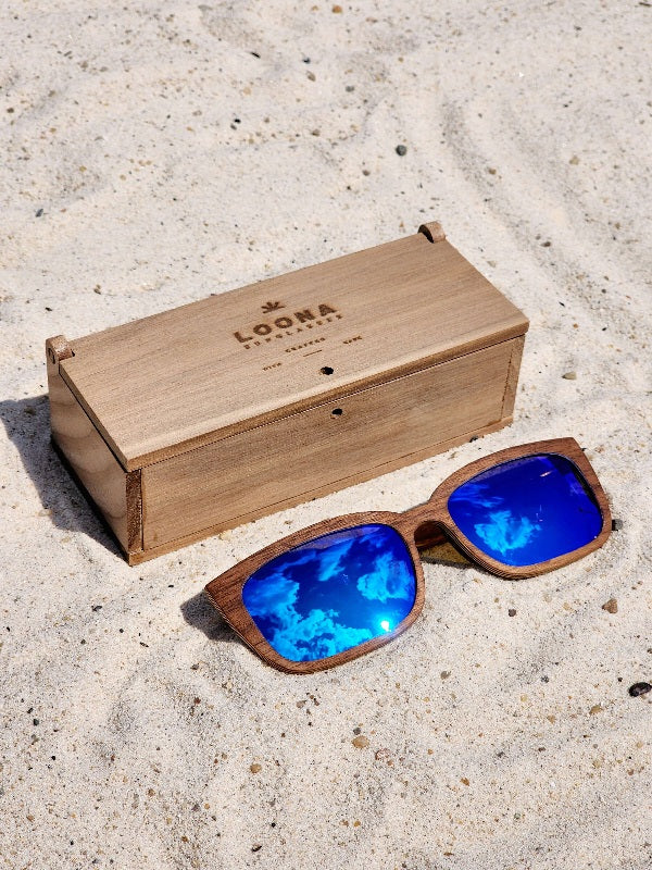 Walnut wooden sunglasses with blue polarized lenses and a box on sandy beach.