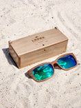 Load image into Gallery viewer, Walnut wooden sunglasses with green polarized lenses and a box on sandy beach.
