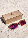 Load image into Gallery viewer, Walnut wooden sunglasses with red polarized lenses and a box on sandy beach.
