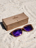 Load image into Gallery viewer, Walnut wooden sunglasses with purple polarized lenses and a box on sandy beach.
