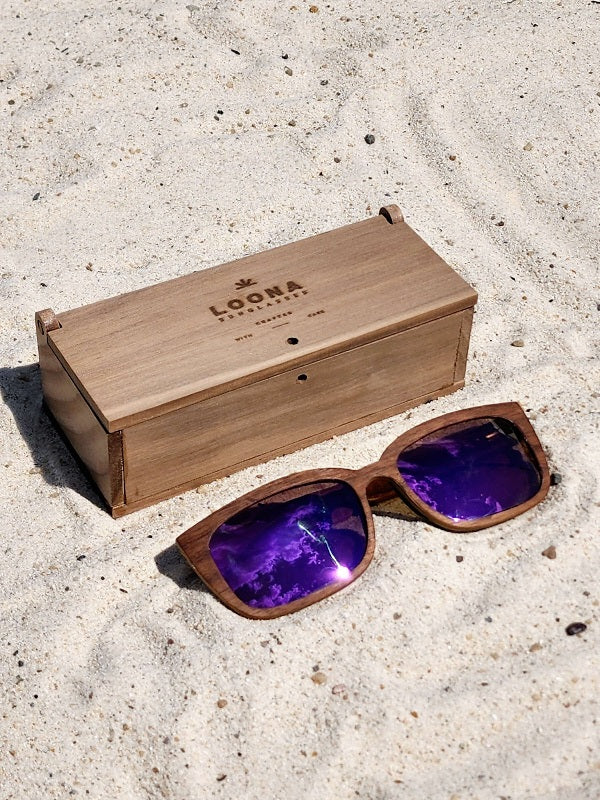 Walnut wooden sunglasses with purple polarized lenses and a box on sandy beach.