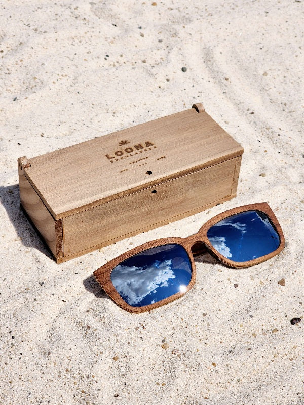 Walnut wooden sunglasses with silver polarized lenses and a box on sandy beach.