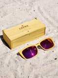 Load image into Gallery viewer, Yellowheart wooden sunglasses with red polarized lenses and a box on sandy beach.
