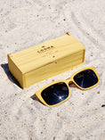 Load image into Gallery viewer, Yellowheart wooden sunglasses with black polarized lenses and a box on sandy beach.
