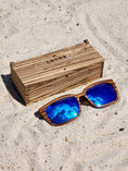 Load image into Gallery viewer, Zebrawood wooden sunglasses with blue polarized lenses and a box on sandy beach.
