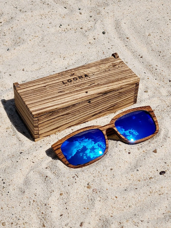 Zebrawood wooden sunglasses with blue polarized lenses and a box on sandy beach.
