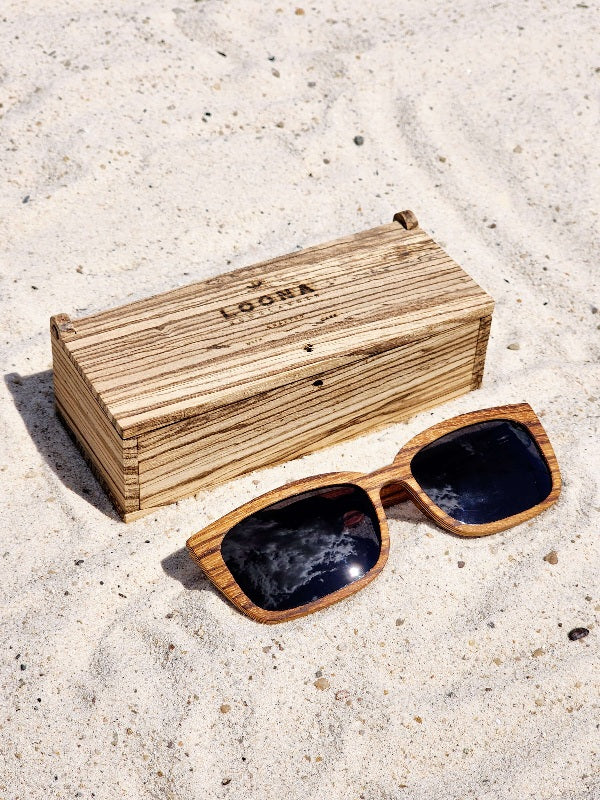 Zebrawood wooden sunglasses with black polarized lenses and a box on sandy beach.