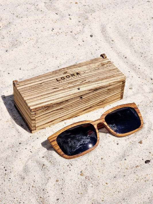 Zebrawood wooden sunglasses with black polarized lenses and a box on sandy beach.