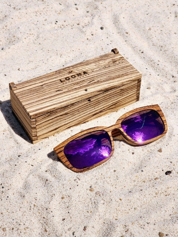 Zebrawood wooden sunglasses with purple polarized lenses and a box on sandy beach.