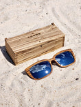 Load image into Gallery viewer, Zebrawood wooden sunglasses with silver polarized lenses and a box on sandy beach.
